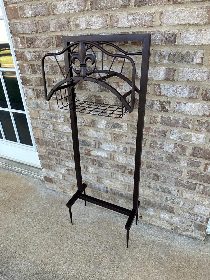Op de afbeelding: Een donkerbruine metalen tuinslanghouder met een fleur-de-lis-ontwerp. De houder heeft een rechthoekig frame, een gebogen bovenkant en een draadmand voor opslag. De houder is ontworpen om rechtop te staan.