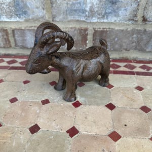 May include: A cast iron goat figurine in a rustic brown finish. The goat has curved horns and is standing on four legs. The figurine is a decorative item, suitable for home decor or as a garden ornament.