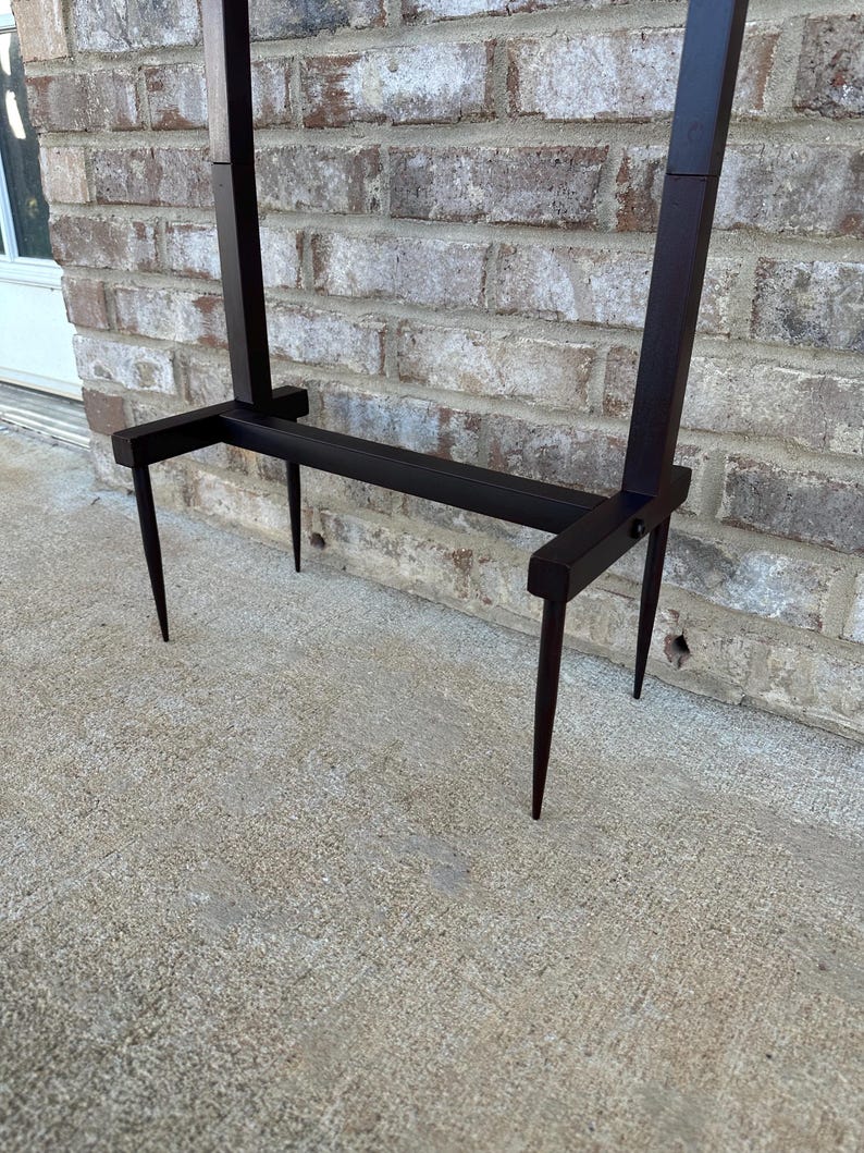 Op de afbeelding: Een donkerbruine metalen plantenstandaard met een rechthoekige basis en twee verticale steunen. De standaard heeft een minimalistisch ontwerp met puntige poten, geschikt voor buiten of binnen. De standaard staat op een betonnen oppervlak voor een bakstenen muur.