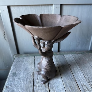 May include: A cast iron rabbit figurine holding a flower-shaped bowl. The bowl is brown and the rabbit is a dark brown color.