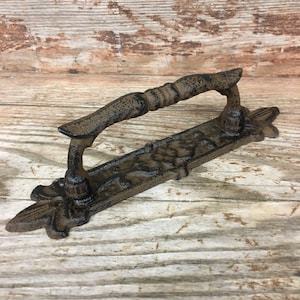May include: A rustic, cast iron door pull with a decorative handle and ornate base. The door pull is finished in a dark brown color, with visible texture and detailing. The handle is approximately 6 inches long.