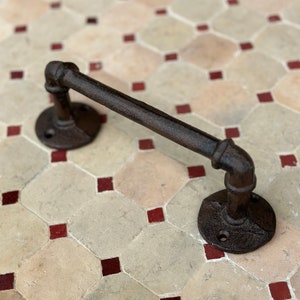 May include: A black metal towel bar with a pipe design. The bar is mounted on a wall with a tiled background.