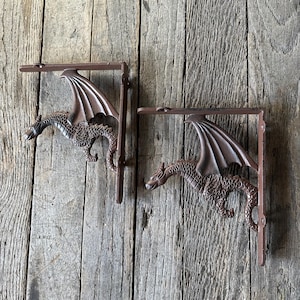 May include: Two brown metal dragon shelf brackets. Each bracket features a detailed dragon with outstretched wings, mounted on a rectangular support. The brackets are designed to be mounted on a wall and are made of cast iron.