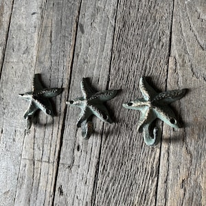 May include: Three bronze-colored starfish-shaped hooks arranged on a weathered wooden surface. Each hook has five points and a curved base for hanging items. The hooks are a muted green-bronze color.