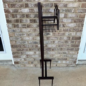 Op de afbeelding: Een hoge, donkerbruine metalen plantenstandaard met een rechthoekige achterkant, een kleine plank en een draadmand. De standaard heeft een decoratief krulontwerp aan de bovenkant en twee poten aan de onderkant. Voor buitengebruik.