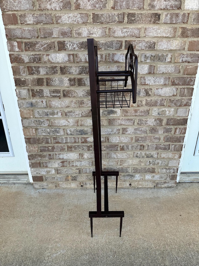 Op de afbeelding: Een hoge, donkerbruine metalen plantenstandaard met een rechthoekige achterkant, een kleine plank en een draadmand. De standaard heeft een decoratief krulontwerp aan de bovenkant en twee poten aan de onderkant. Voor buitengebruik.