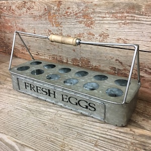 May include: A galvanized metal egg holder with a wooden handle and the words "FRESH EGGS" printed on the front. The holder has 12 circular compartments for eggs and a metal handle for carrying.