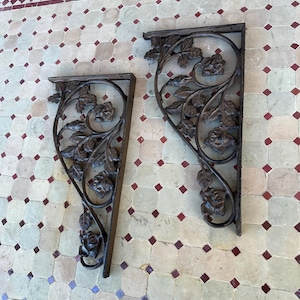 May include: Two ornate, dark metal shelf brackets. Each bracket features a floral design with roses and leaves. The brackets are positioned on a tiled surface with a mix of neutral and red-toned tiles, creating a vintage aesthetic.