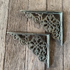 May include: Two ornate, cast iron shelf brackets with a distressed, aged finish. The brackets feature a decorative floral and scroll design, perfect for adding a vintage touch to any space. The brackets are shown against a rustic wooden background.
