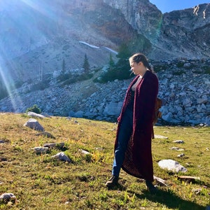 May include: A person wearing a long, maroon cardigan in a mountainous landscape. The sun is shining brightly and there are rocks and grass in the background.