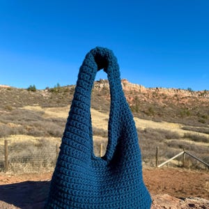 Könnte beinhalten: Eine türkisfarbene gehäkelte Tragetasche mit einem einzigen Riemen. Die Tasche liegt auf einem großen Felsen.