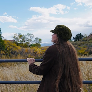 May include: A woman wearing a brown jacket and a green newsboy cap looks out over a field of tall grass. The field is bordered by a black metal fence.