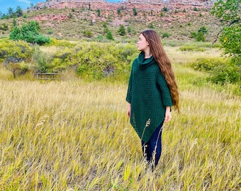 Op de afbeelding: Een vrouw draagt een donkergroene gehaakte poncho met een rolkraag en staat in een veld met hoog gras.