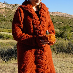 Puede incluir: Un cárdigan largo de crochet naranja con capucha y un patrón de punto de cable. El cárdigan lo lleva puesto una persona que sostiene una pequeña calabaza de crochet naranja.