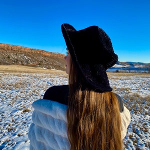 May include: A person wearing a black wide-brimmed hat and a white faux fur jacket stands in a snowy field, looking out at a distant mountain range.