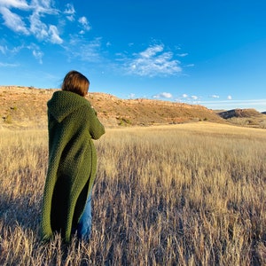 Pode incluir: Uma pessoa vestindo um longo cardigã verde escuro com capuz está em um campo de grama alta e seca. A pessoa está olhando para uma colina distante.