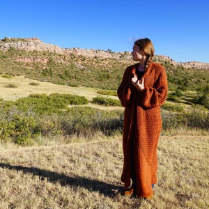 Puede incluir: Una mujer con un largo cárdigan de punto color óxido en un campo con una cordillera de montañas al fondo.