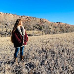 Puede incluir: Una mujer con una chaqueta de sherpa marrón con cuello de piel sintética marrón está de pie en un campo de hierba seca. Está mirando hacia un lado, y hay árboles y una ladera rocosa en el fondo.