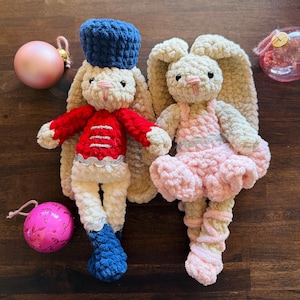 May include: Two handmade crochet bunny dolls. One bunny wears a red jacket, blue hat, and white pants. The other bunny is dressed in a pink dress. Both have long ears and are cream-colored. Christmas ornaments are in the background.