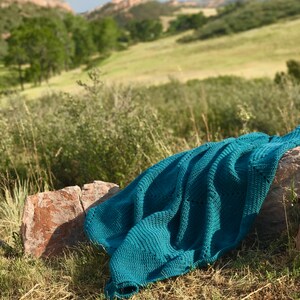 May include: A teal crocheted blanket draped over a rock in a grassy field with a blurred background of rolling hills.
