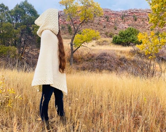 May include: A cream-colored, hooded crochet poncho. The hood has a textured, raised pattern. The poncho is worn over dark pants and is set against a backdrop of a field of tall, dry grass, trees, and a mountain in the distance.