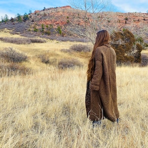 May include: A long, brown crocheted cardigan with an open front and a loose fit. The cardigan is being worn by a person standing in a field of long grass.