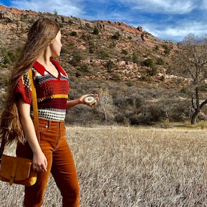 Op de afbeelding: Een vrouw in een kleurrijk gestreept gehaakt topje en een bruine corduroy broek staat in een veld en houdt een kompas vast. Ze kijkt naar een rotsachtige heuvel in de verte.