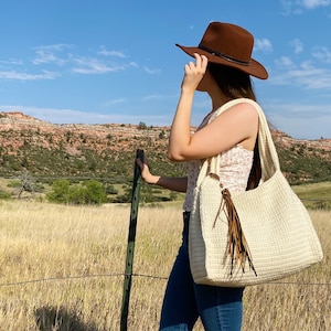 May include: A woman wearing a brown cowboy hat and blue jeans is holding a white crocheted tote bag with a brown leather strap and fringe. The bag is large and spacious, perfect for carrying everyday essentials.