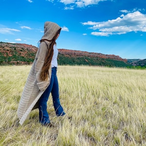 Pode incluir: Um casaco longo de crochê bege com capuz, usado por uma pessoa em pé em um campo de grama alta. A pessoa está vestindo jeans azul e uma camiseta branca.