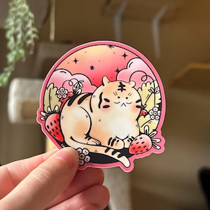May include: A pink and orange sticker with a cartoon tiger sitting on a bed of strawberries and flowers. The tiger has a pink nose and a white stripe on its forehead. The background is a pink and orange gradient with white clouds.