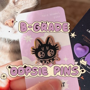 Può includere: Primo piano di una carta rosa e bianca con il testo "B-GRADE OOPSIE PINS" e uno spillo in smalto nero e oro di un gatto con un collare a punte. La carta ha anche il testo "hard enamel pins" e una piccola stella.