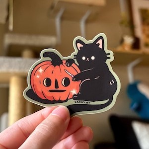 May include: A black cat with white eyes is carving a jack-o'-lantern with a knife. The pumpkin is orange and has a sparkly face. The sticker says "CAMCCAT".
