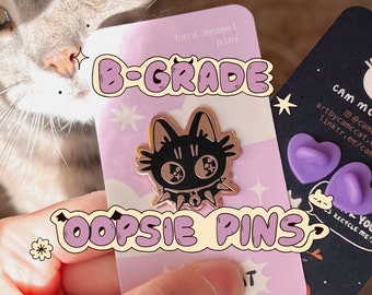 May include: A close-up of a pink and white card with the text "B-GRADE OOPSIE PINS" and a black and gold enamel pin of a cat with a spiked collar. The card also has the text "hard enamel pins" and a small star.
