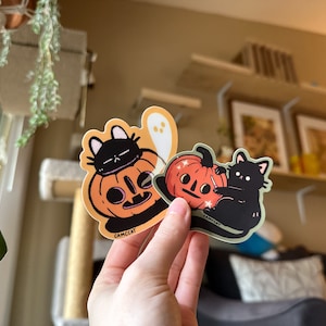 Pode incluir: Dois adesivos de Halloween com gatos pretos e ab&oacute;boras. Um adesivo mostra um gato preto usando uma ab&oacute;bora na cabe&ccedil;a. O outro adesivo mostra um gato preto esculpindo uma ab&oacute;bora com uma faca. Ambos os adesivos t&ecirc;m o texto "CAMCCAT" neles.