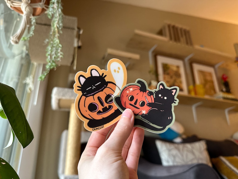 Pode incluir: Dois adesivos de Halloween com gatos pretos e ab&oacute;boras. Um adesivo mostra um gato preto usando uma ab&oacute;bora na cabe&ccedil;a. O outro adesivo mostra um gato preto esculpindo uma ab&oacute;bora com uma faca. Ambos os adesivos t&ecirc;m o texto "CAMCCAT" neles.