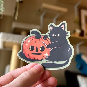 Pode incluir: Um gato preto com olhos brancos est&aacute; a esculpir uma ab&oacute;bora de Halloween com uma faca pequena. A ab&oacute;bora tem uma cara amig&aacute;vel e brilhos &agrave; sua volta. O autocolante diz "CAMCCAT" na parte inferior.