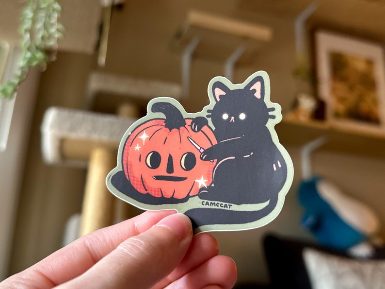 Pode incluir: Um gato preto com olhos brancos est&aacute; a esculpir uma ab&oacute;bora de Halloween com uma faca pequena. A ab&oacute;bora tem uma cara amig&aacute;vel e brilhos &agrave; sua volta. O autocolante diz "CAMCCAT" na parte inferior.