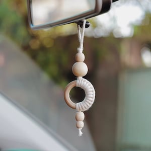 Może przedstawiać: Biały wisiorek do samochodu z makramą i drewnianymi koralikami. Wisiorek jest zawieszony na lusterku wstecznym samochodu.