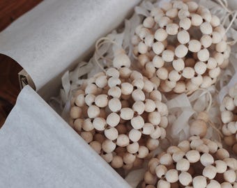 May include: A collection of spherical ornaments made of small, light-colored wooden beads. The ornaments are nestled in a box lined with white tissue paper. The beads are strung together to form a ball shape, and each has a loop for hanging.