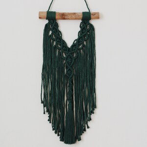 May include: A dark green macrame wall hanging with a wooden dowel at the top. The hanging features a V-shaped design with fringe details.