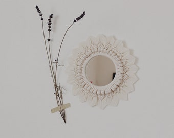 May include: A white macrame mirror with a round mirror in the center. The mirror is hanging on a white wall with dried lavender flowers to the left.