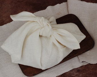 Puede incluir: Una bolsa de tela blanca atada con un nudo, sentada sobre una tabla de madera y una superficie de tela beige.
