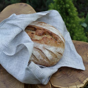 Puede incluir: Un pan redondo con corteza dorada y cortes, envuelto en un paño de lino a rayas. El paño presenta finas rayas grises. La escena está ambientada sobre una superficie de madera con forma natural y orgánica.