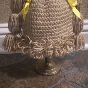 May include: A crocheted doll hat with brown yarn and yellow ribbon pigtails. The hat has a crocheted ruffle around the bottom and is displayed on a gold stand.
