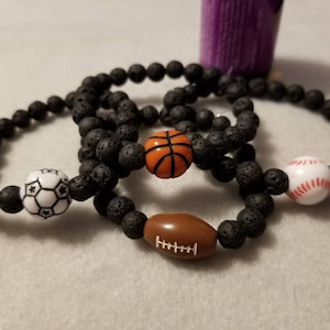 May include: Three black lava rock bracelets with different sports ball charms. The charms are a soccer ball, a basketball, and a baseball.