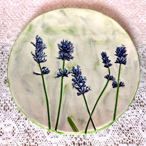 May include: A round ceramic plate with a white background and a green and white marbled design. The plate features a hand-painted design of lavender flowers in shades of blue and purple.