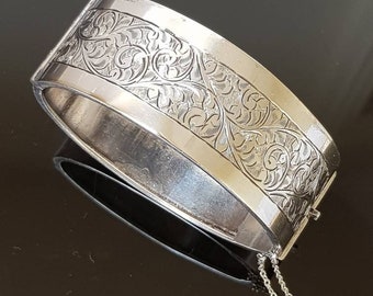 May include: A silver bangle bracelet with an intricate floral design engraved on the surface. The bracelet has a hinged clasp and a delicate chain attached.