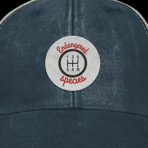 May include: A blue and beige trucker hat with a distressed look. The hat features a round patch with a white gear shift pattern and the text "Endangered Species".