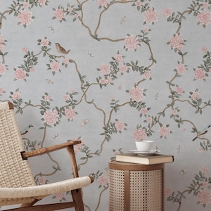 May include: A light blue floral wallpaper featuring pink roses, brown branches, and small birds. A woven chair, a small side table, and books are in the foreground. The wallpaper creates a serene and natural ambiance.