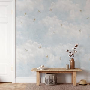 May include: A light blue and white cloud-themed wallpaper with gold bird accents. A wooden bench with a woven top, a brown vase with cotton stems, and a woven basket are in front of the wall. A straw hat sits on the floor.
